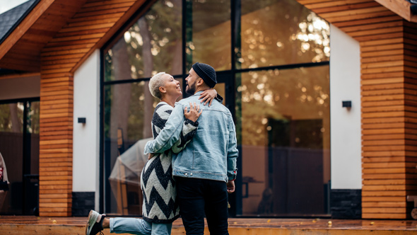 couple outside modern wooden house, large glass windows, embracing happily